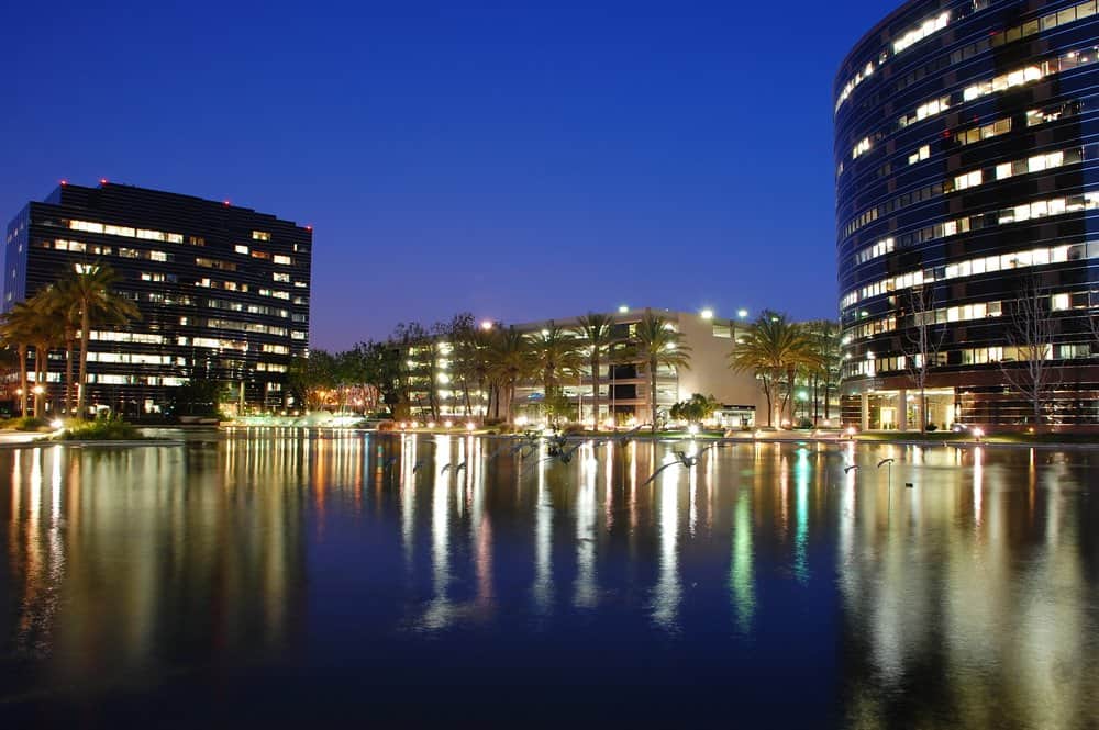 Night view of modern city buildings reflected in water with palm trees.