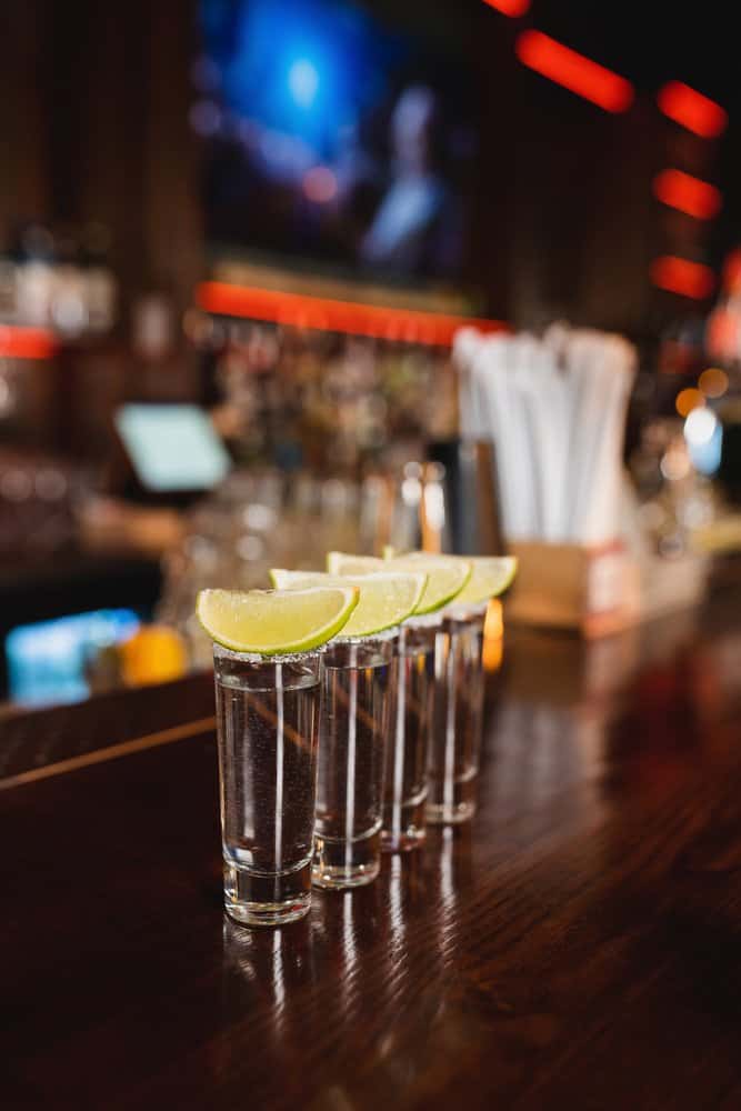 Four tequila shots with lime wedges on a bar counter at a lively bar.