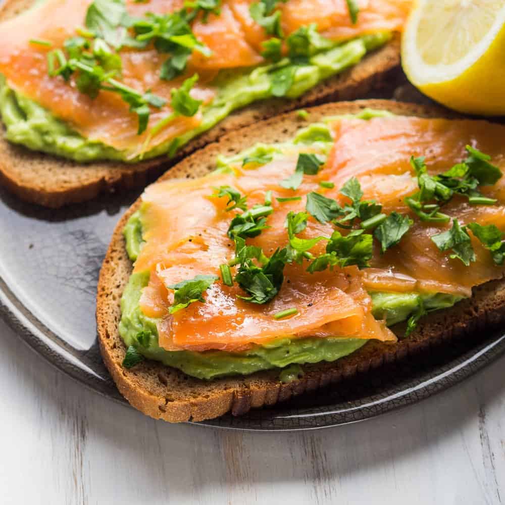 Smoked salmon avocado toast on whole grain bread with lemon wedge.